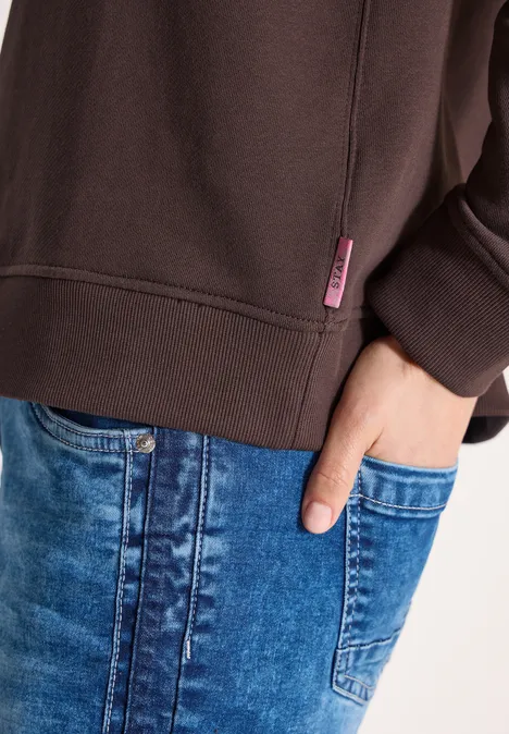 Oberkörper mit braunem Sweatshirt und mit Hand in die Tasche gesteckter Hand über blauen Jeans
